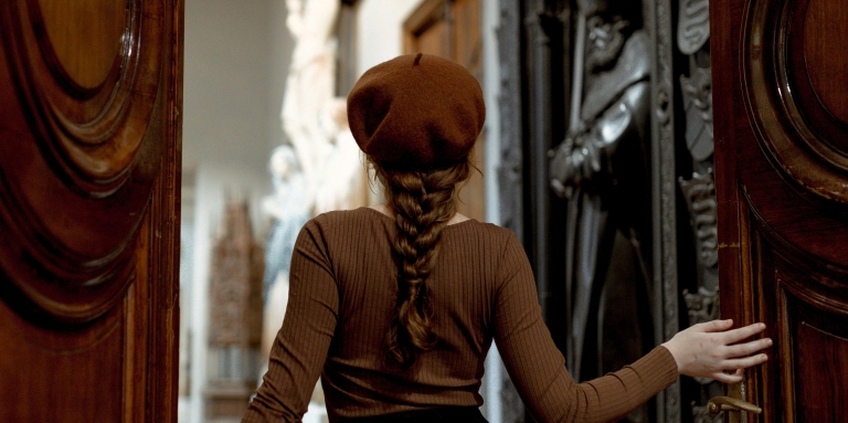 Une femme coiffée d’un béret brun entre dans une salle de musée ornée de sculptures en poussant une grande porte en bois.