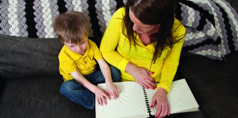 A little boy sits next to his mother on a couch reading a tactile book. Both are in yellow shirts.