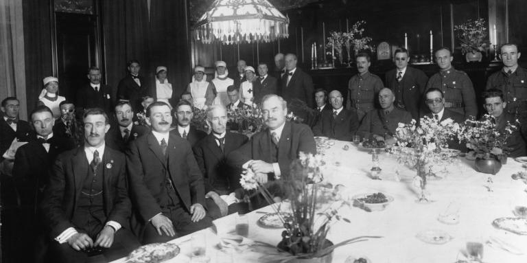 A group of men in suits sitting in a room with a dining table in from of them.