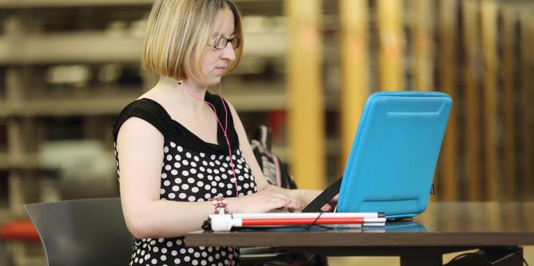 Une femme est assise à un bureau avec son ordinateur portable ouvert et sa canne blanche effondrée à côté.