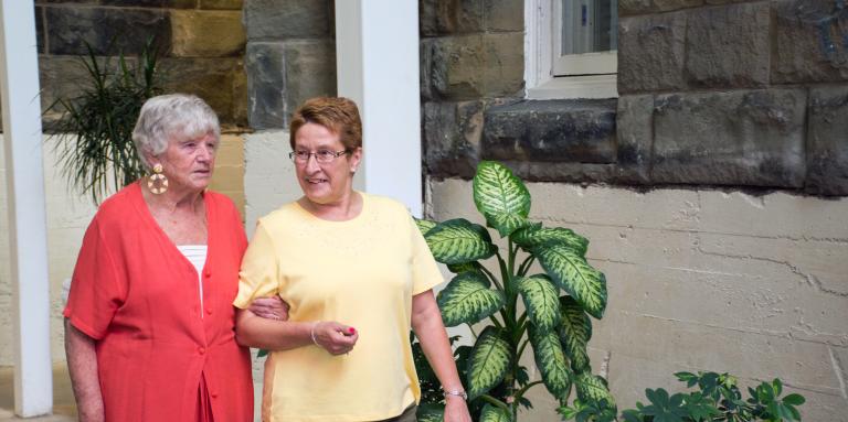 Elderly woman in red is walking and being guided by another woman who is wearing a yellow shirt and olive green pants.