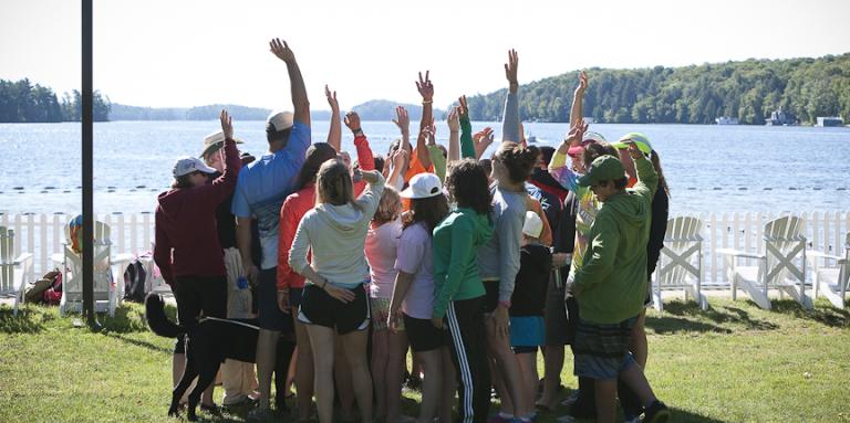 Groupe de jeunes participants du camp, devant un lac, levant les bras en l’air fièrement.