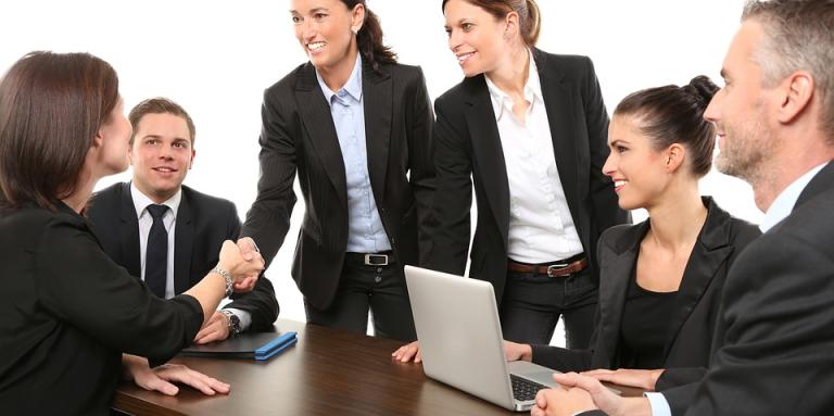 Two women shake hands over a table of six business people.