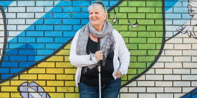 Wendy, holding her white cane, stands in front of a brick wall with a colourful mural.