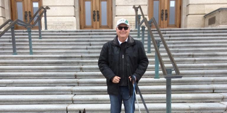 Bob Huber se tient devant l'édifice de l'Assemblée législative de la Saskatchewan en souriant et portant ses lunettes de soleil sur sa casquette des Roughrider.