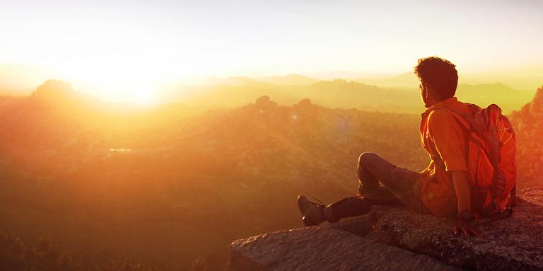 A man sits on the edge of a cliff, looking at the sun rise.
