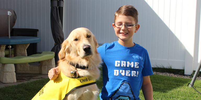 Un garçon souriant portant des lunettes avec son bras autour d'un Golden Retriever qui porte une veste jaune.