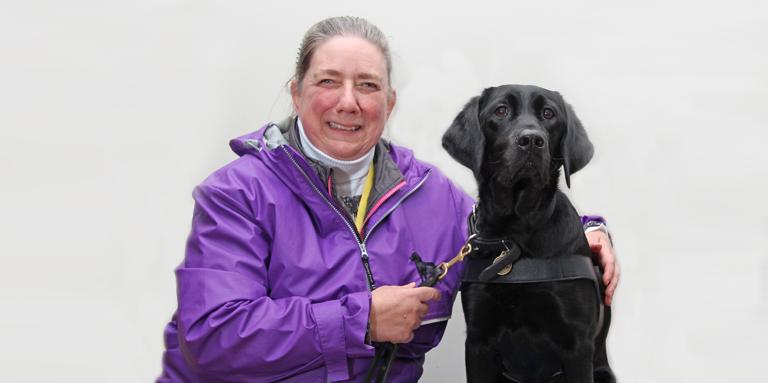 Une femme portant une veste mauve et son chien de race croisée Labrador noir-Golden Retriever chien-guide.