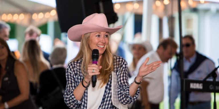 Nancy Simonot, portant un chapeau de cowgirl rose et une chemise à carreaux, un micro à la main, s'adresse à une foule. 