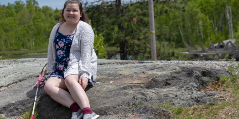 Alicia smiles and sits outdoors on a large rock. Behind her are trees. She is wearing a summer floral dress. Her white cane rests on the rock to the right of her.