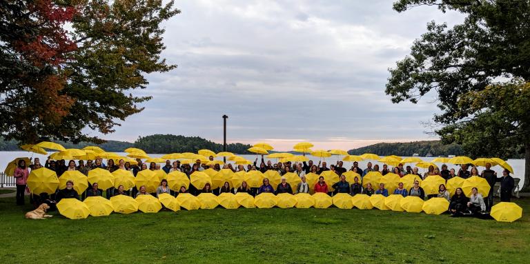 Un grand groupe de plus de 50 employés d’INCA tenant des parapluies jaunes d’INCA sur les rives du Centre Lake Joseph d’INCA.  