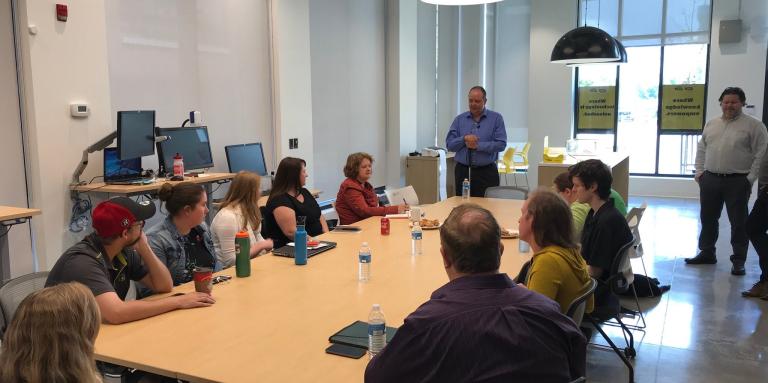 A large table surrounded with seated participants. Wayne Henshall stands at the front of the room with his white cane and addresses the participants.
