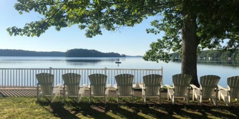 Une vue du lac par une journée ensoleillée avec une rangée de chaises Muskoka blanches au bord de la plage.