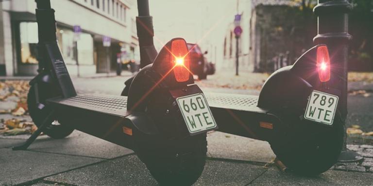 Two e-scooters sit side-by-side on a sidewalk. Their break lights are blinking red.