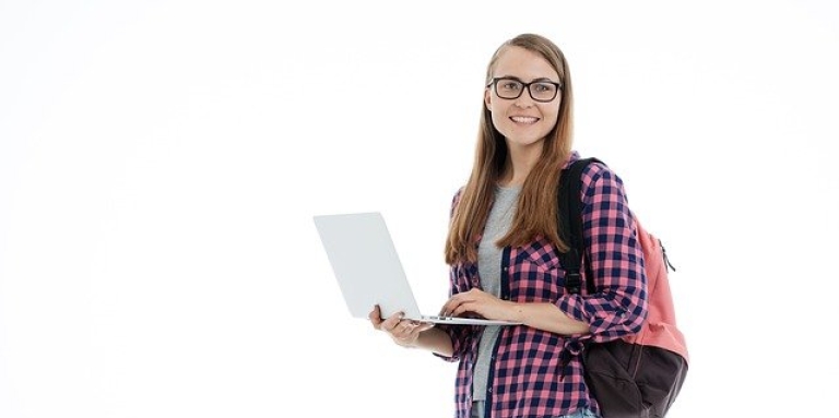 Une collégienne debout tient un ordinateur portable et tape de sa main gauche. Elle porte un sac à dos.