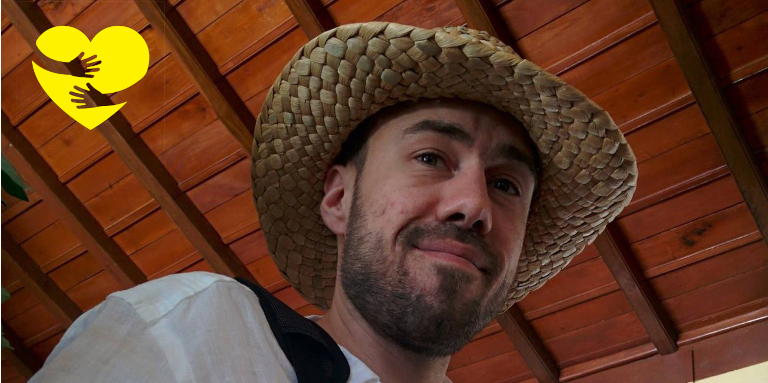 Kevin, looking down at the camera with a smile while wearing a straw hat. A graphic of arms hugging a yellow cartoon heart can be seen in the top-left corner of the photo.
