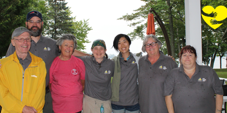 Sandra, wearing a pink shirt, smiling for the camera in a group photo with other CNIB Lake Joe volunteers. A graphic of arms hugging a cartoon yellow heart can be seen in the top-right corner of the photo.