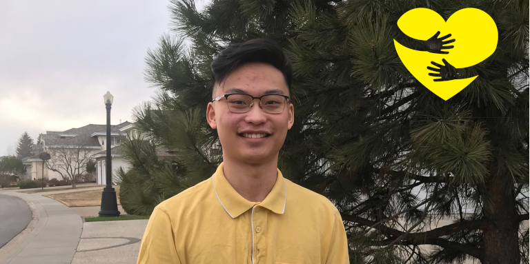 Warren, standing outside wearing a yellow shirt, smiling for the camera. A graphic of arms hugging a yellow cartoon heart can be seen in the top-right corner of the photo.