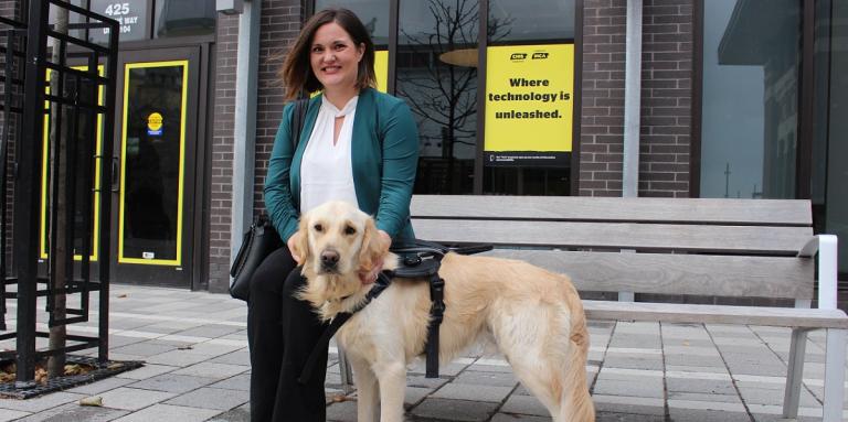 A smiling woman sitting on a bench with her CNIB Guide Dog, outside a CNIB building with yellow branded CNIB signs and posters.