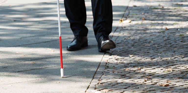  A white cane scans a sidewalk. In the background, mens shoes trail behind the tip of the cane.