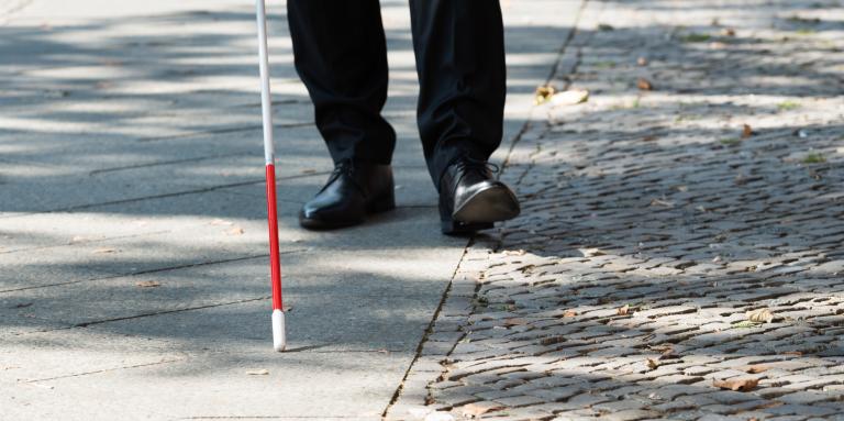 A white cane scans a sidewalk. In the background, mens shoes trail behind the tip of the cane.