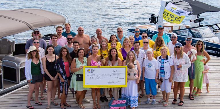 A group shot of the partygoers at the Dock to Dock event. Together they hold a large cheque. 