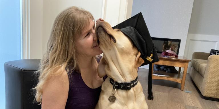 Laurie sits on her living room floor with her guide dog, Bridget, a yellow Labrador retriever-golden retriever cross. Bridget wears a graduation cap and sits in Laurie’s lap. Laurie affectionately presses her face against the Bridget’s fluffly face. 