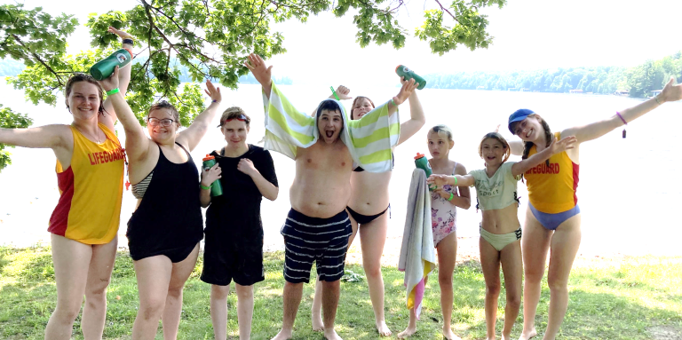 a group of campers stand in front of the waterfront with their bathing suits on. They are throwing their hands up in the air and have big smiles. Two lifeguards stand beside them. 