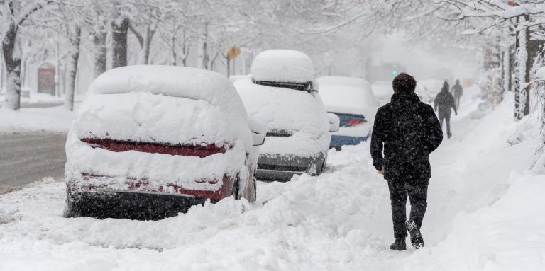 A residential street, sidewalks, and parked cars are covered in snow. A man walks down a snow-covered sidewalk.