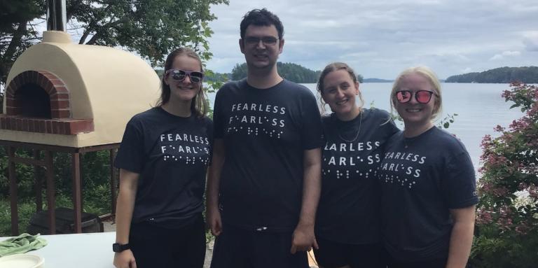 From left to right: (L-R) Emilee Schevers, Tavish Gillan, Taylor Gaudon and Jade Ondrik by the outdoor pizza oven at CNIB Lake Joe. They are wearing blue t-shirts with the word “Fearless” in print and braille.