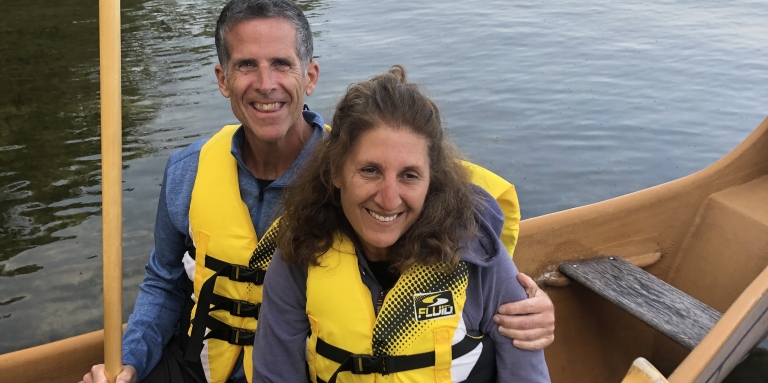 Matt et Miriam sont assis côte à côte dans un canoë se trouvant sur l'eau. Ils portent des shorts et un gilet de sauvetage jaune, et tiennent une pagaie en bois. Des arbres se profilent à l’horizon et le ciel est bleu.