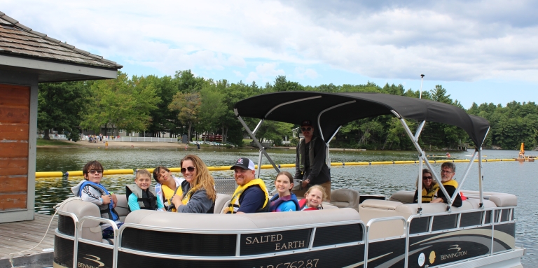 A group of smiling CNIB Lake Joes campers on a pontoon boat near the boathouse.