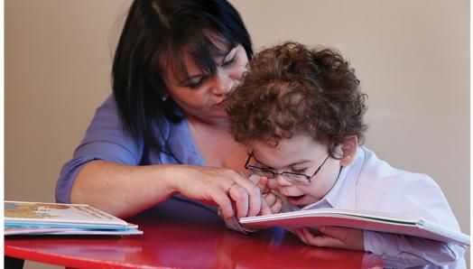 A young boy reads a tactile book. His mother helps guide his hands over the book.