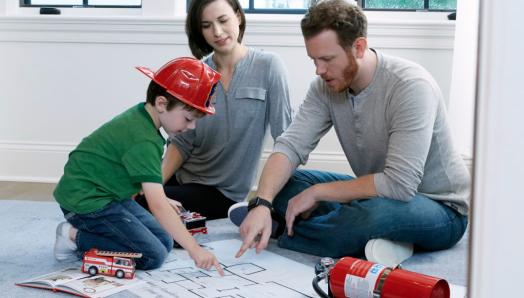 A family sits on the floor looking at a floor plan. There is a red fire extinguisher on the floor and the boy is wearing a firefighter hat.