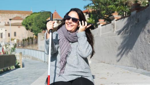 A young woman sits alone outdoors. In her left hand, she is holding her smartphone up to her ear. In her right hand, she holds her white cane