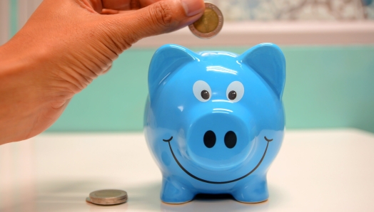 Une main, à gauche, tient un pièce de monnaie au-dessus d'une tirelire bleue.