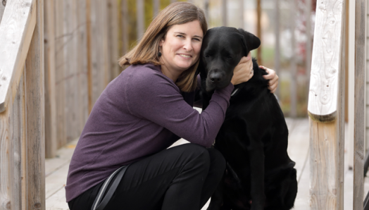 Photo of Shelley sitting on a wooden porch, smiling while giving Rookie a big hug.