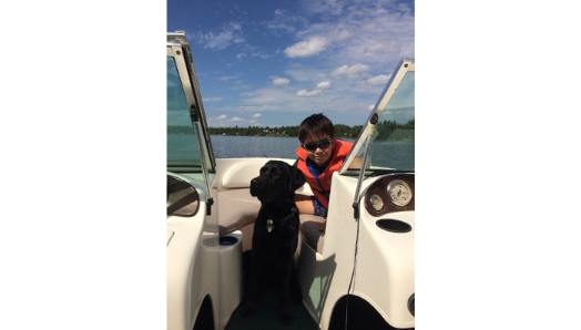 Keaton is sitting on a boat in the water with his arm around Indy, a black Labrador-golden-retriever, who is sitting on the boat beside him.