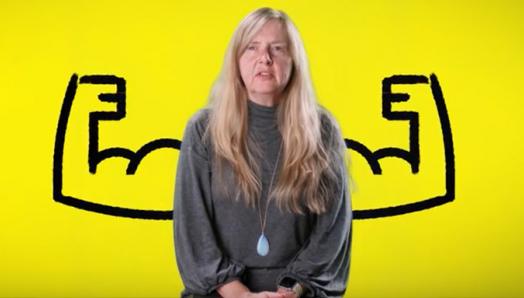 Diane Bergeron se trouve devant un arrière-fond jaune duquel se détache une illustration caricaturale de bras en flexion chaque côté d’elle.