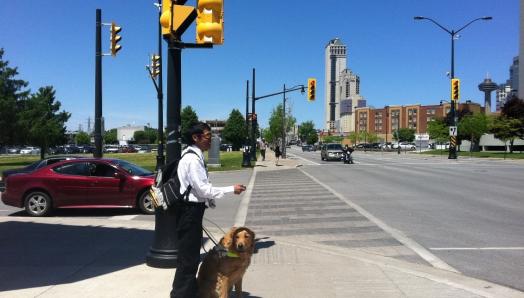 Un jeune homme et son chien guide se tiennent au coin d'un carrefour très fréquenté. Dans sa main droite, il tient une télécommande Key2Access. Dans sa main gauche, il tient le harnais du chien-guide