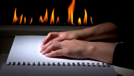 Des mains se déplacent sur un livre en braille. Une cheminée émet des flammes en arrière-plan.