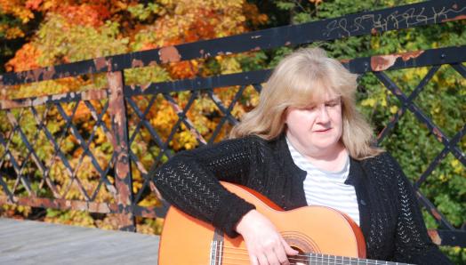 Photo de Christine en train de jouant la guitare sur un pont. Il y a des arbres derrière elle et le soleil brille.
