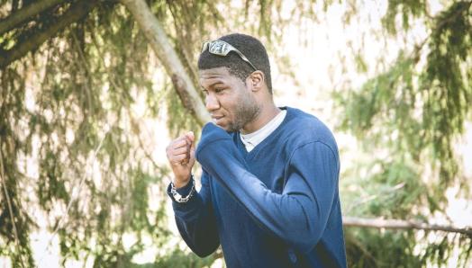 Dressed casually in jeans and a blue shirt, Bernard, strikes a boxing pose. His fists are up near his chest ready to punch. Behind him is a landscape of sweeping green trees. 