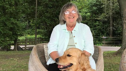 Photo of Claire sitting in a chair outdoors and smiling for the camera. A golden retriever is sitting at her feet.