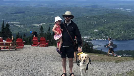 David avec sa fille dans un bras et de l’autre main, tenant dans son harnais son chien-guide Lilo, souriant pour la caméra, le Versant Sud du Mont Tremblant au Québec en arrière-plan