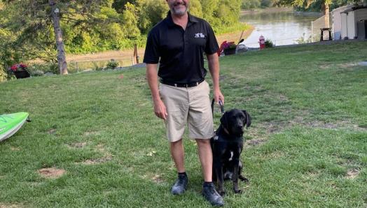 Tracy and his guide dog, Marion, standing on grass in front of a lake on a sunny day, smiling for the camera.