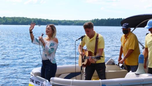 Leah Daniels singing and waving with Will Hebbes beside her playing guitar on a party pontoon, entertaining docks guests on Lake Muskoka.
