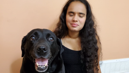 Runa Patel, agenouillée sur le sol auprès de son chien-guide, souriant pour la caméra.