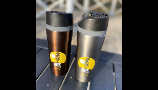 Bronze and charcoal coloured stainless steel travel mugs, with black lids on a table at camp.