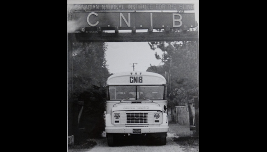 Une photo en noir et blanc d'un ancien autobus scolaire bien marqué CNIB  à l'avant, passant sous le panneau de bienvenue au Centre Lake Joseph d’INCA. 